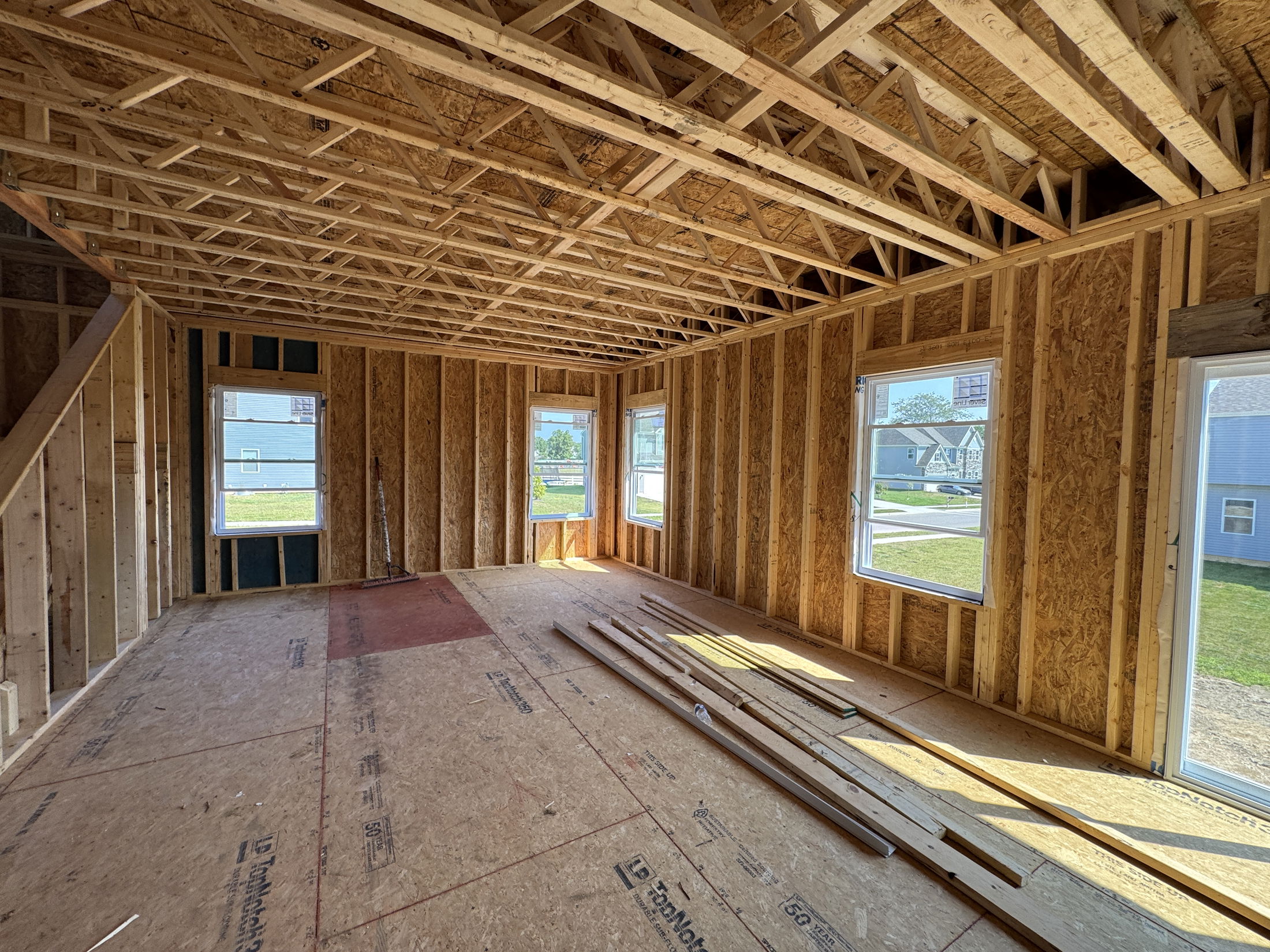 Interior view of a partially constructed house with exposed wooden framing and multiple windows displaying outdoor scenery.