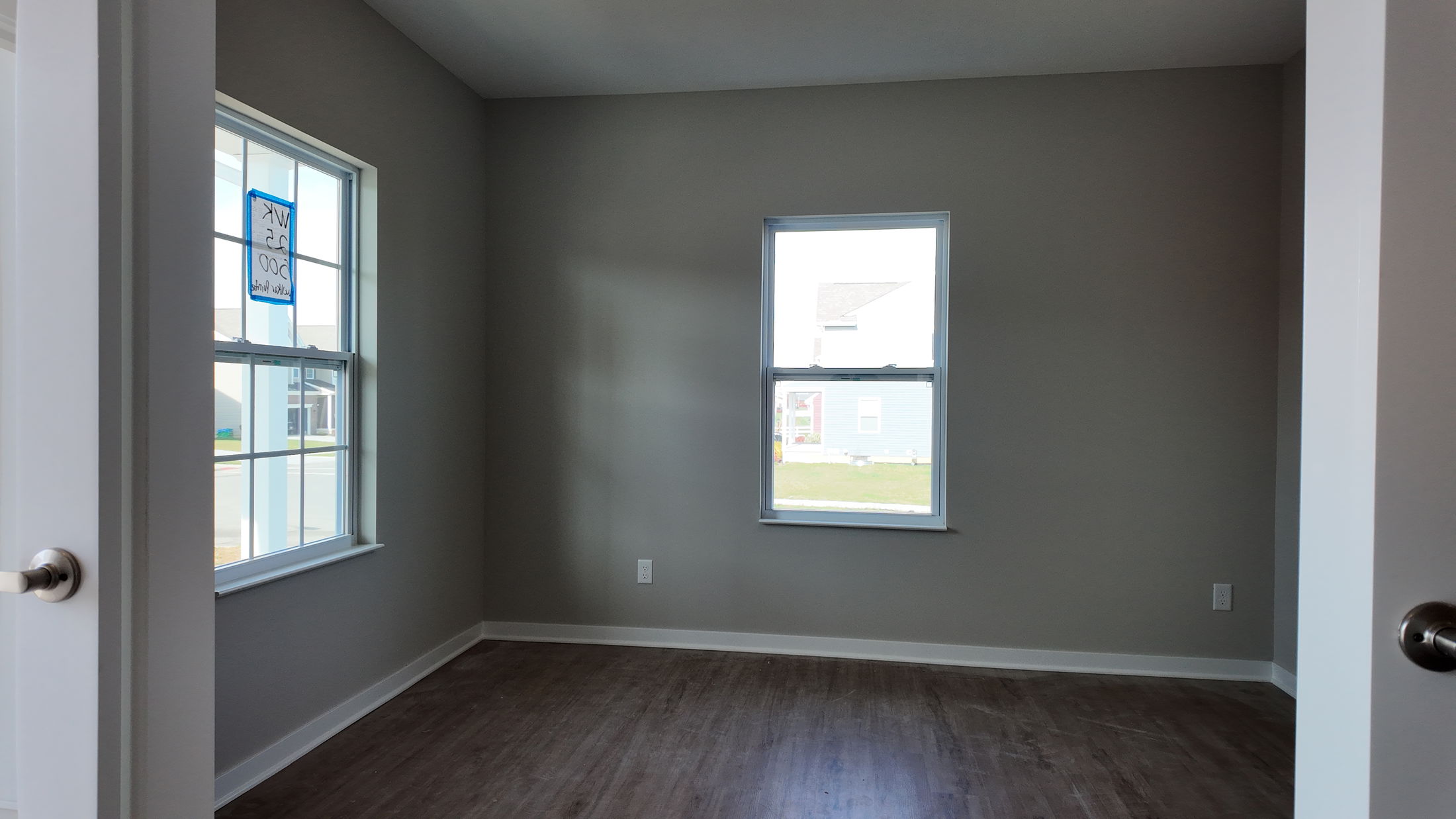 Newly constructed empty room with gray walls, two large windows, and wood flooring.