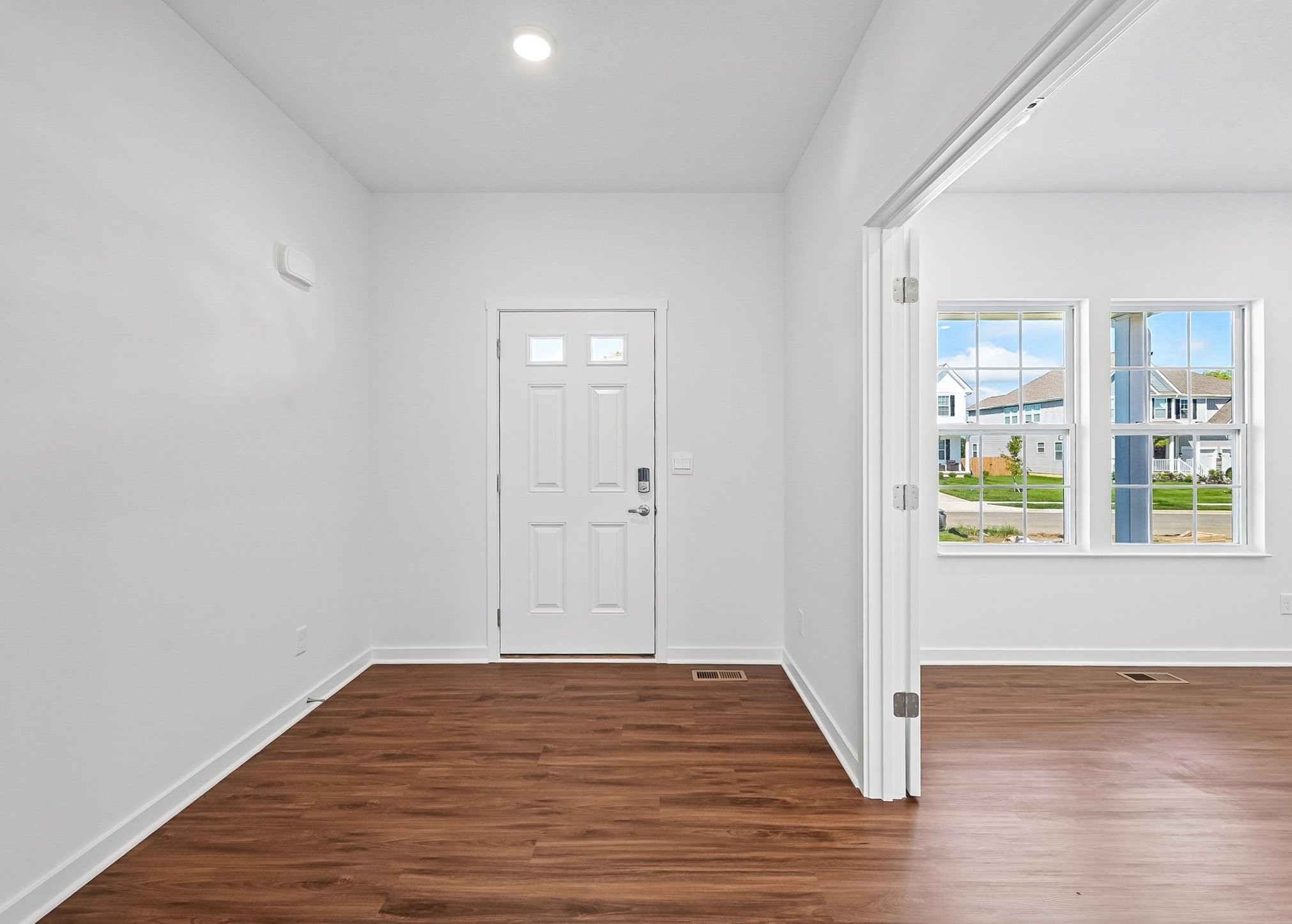 Bright, empty entryway with a front door and large windows showing a view of houses outside.