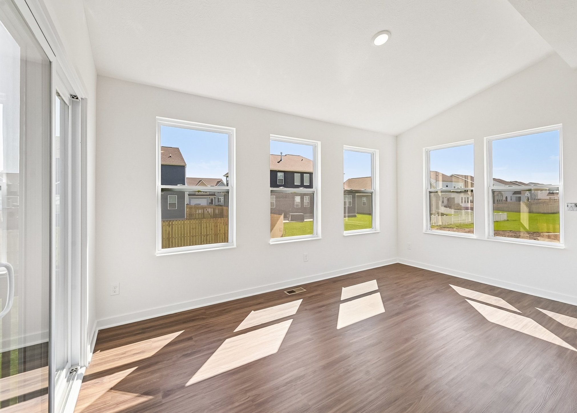 Bright, empty room with large windows and wooden flooring, showcasing a view of green grass and houses outside.