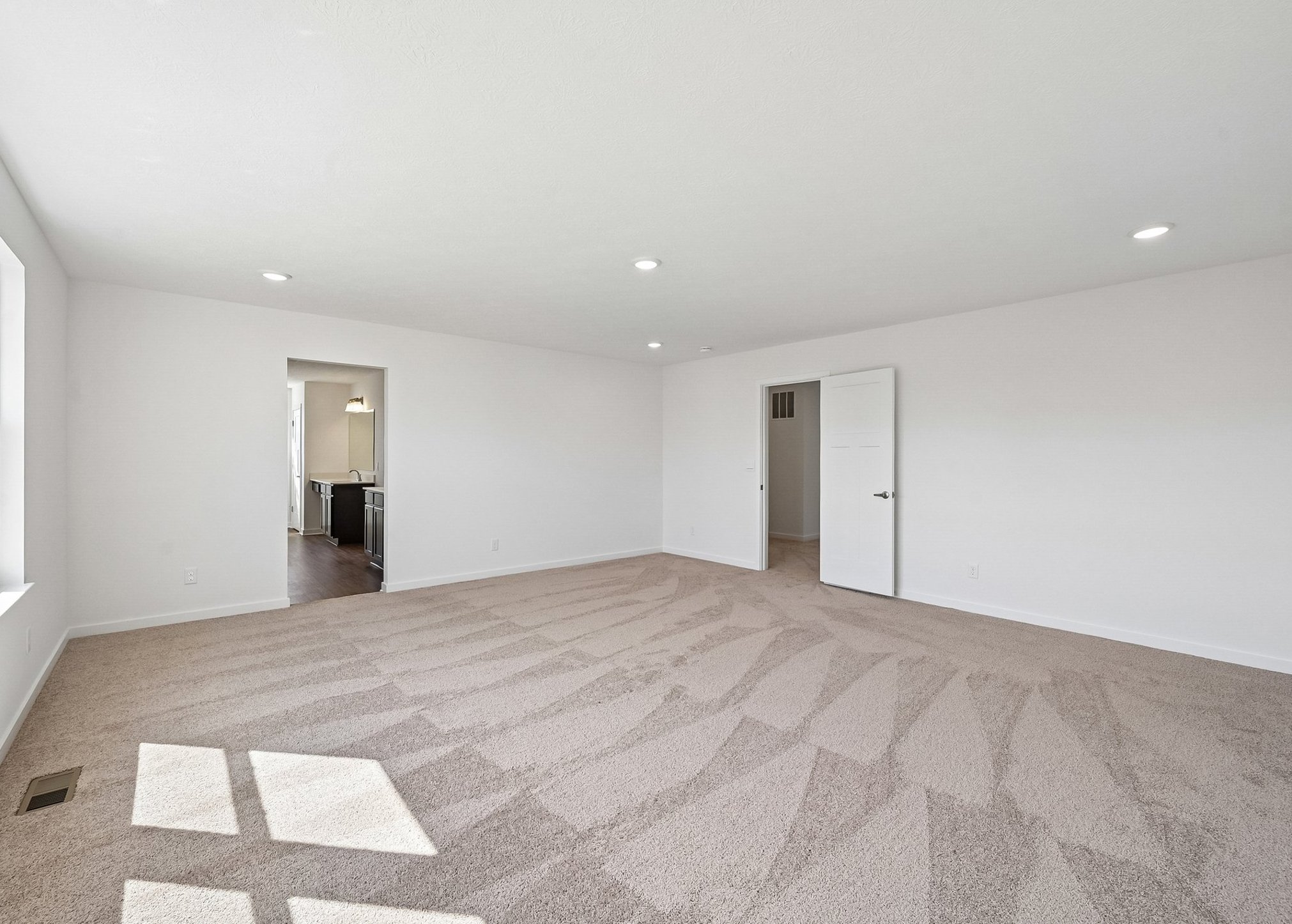 Spacious, empty living room with light gray carpeting and white walls, featuring large windows and recessed lighting.