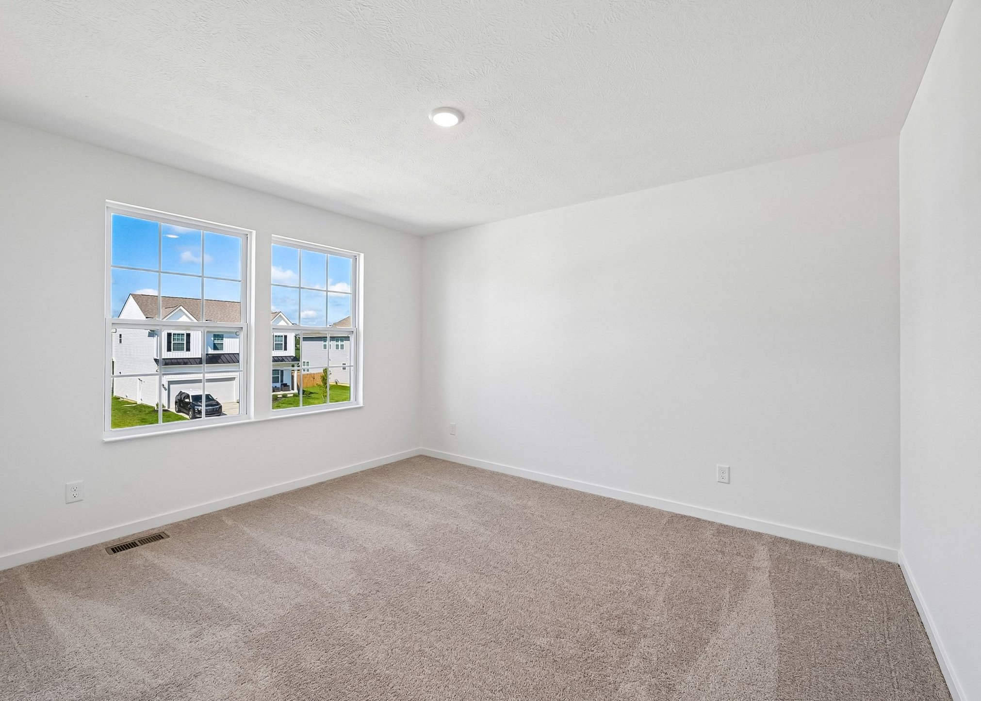 Bright, empty room with two large windows showing an exterior view and light-colored carpet.