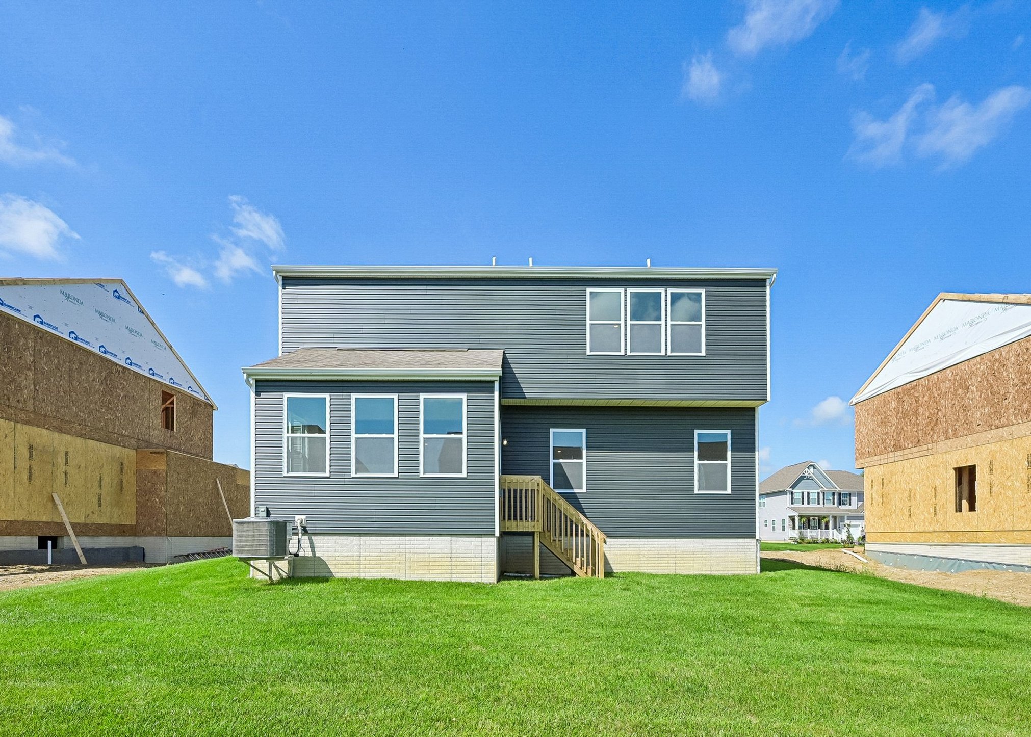 Newly constructed gray two-story house with a wooden staircase and large windows, surrounded by grassy land and neighboring homes under construction.