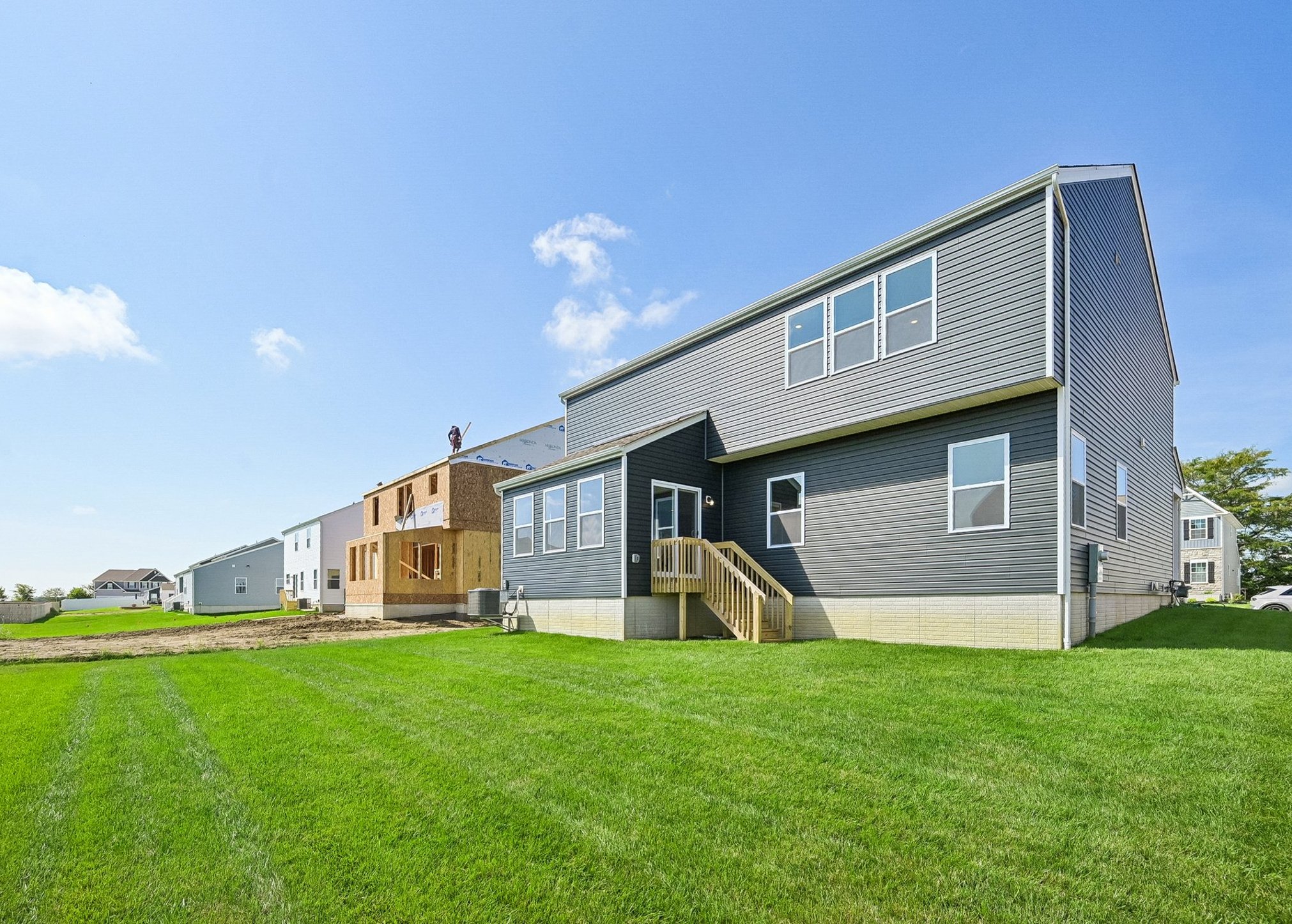 A modern two-story gray house with a wooden deck and green lawn, adjacent to a home under construction.