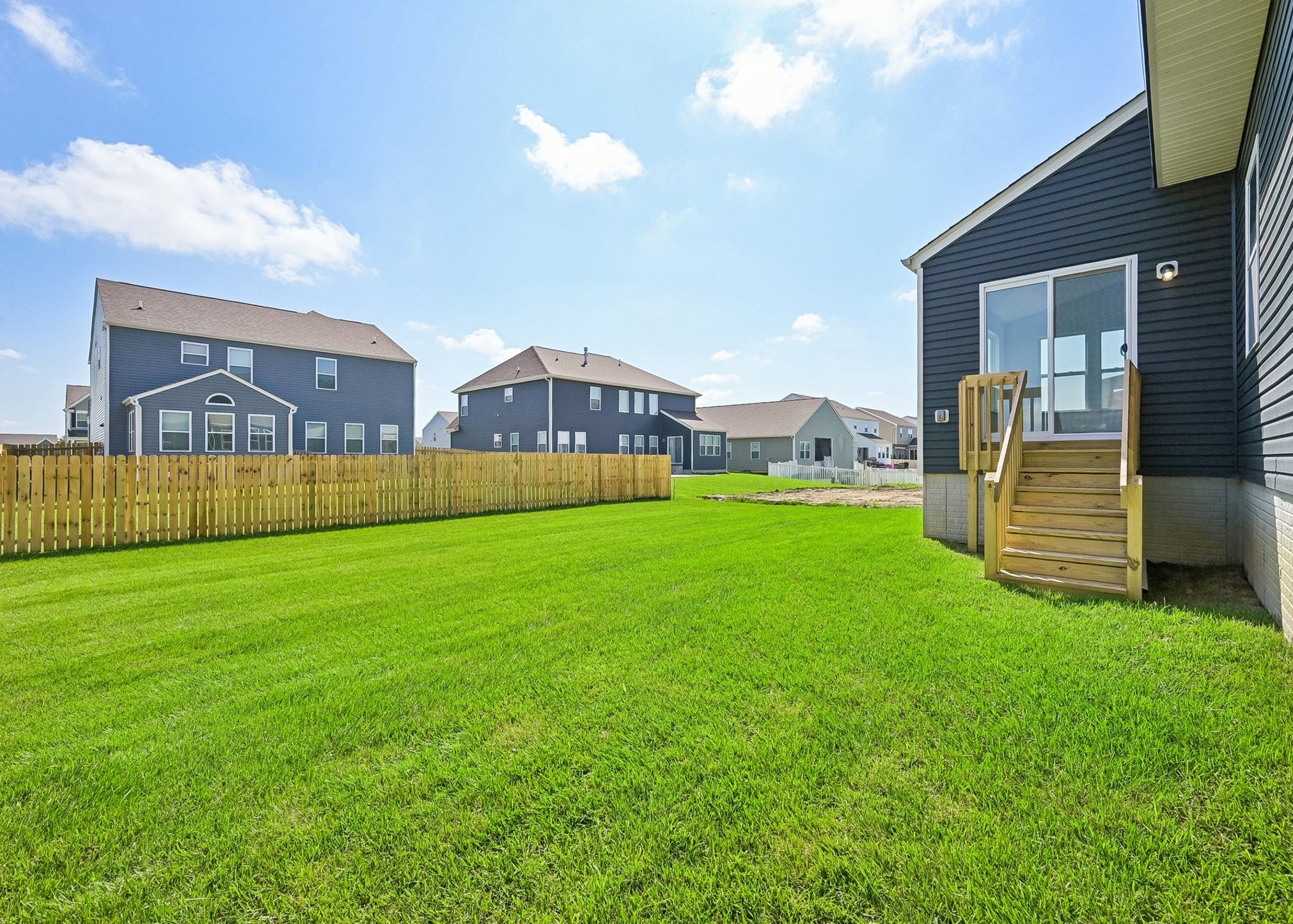 A spacious backyard with green grass and a wooden fence, featuring houses in the background under a clear blue sky.