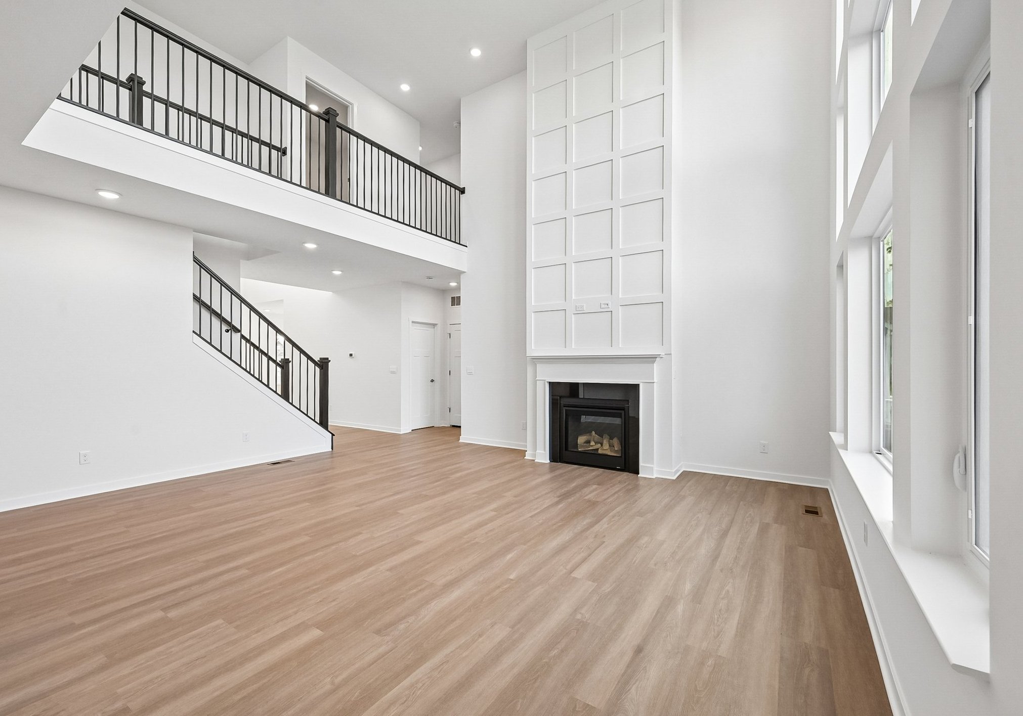 A spacious, modern living room featuring large windows, a fireplace, and a staircase with black railings.