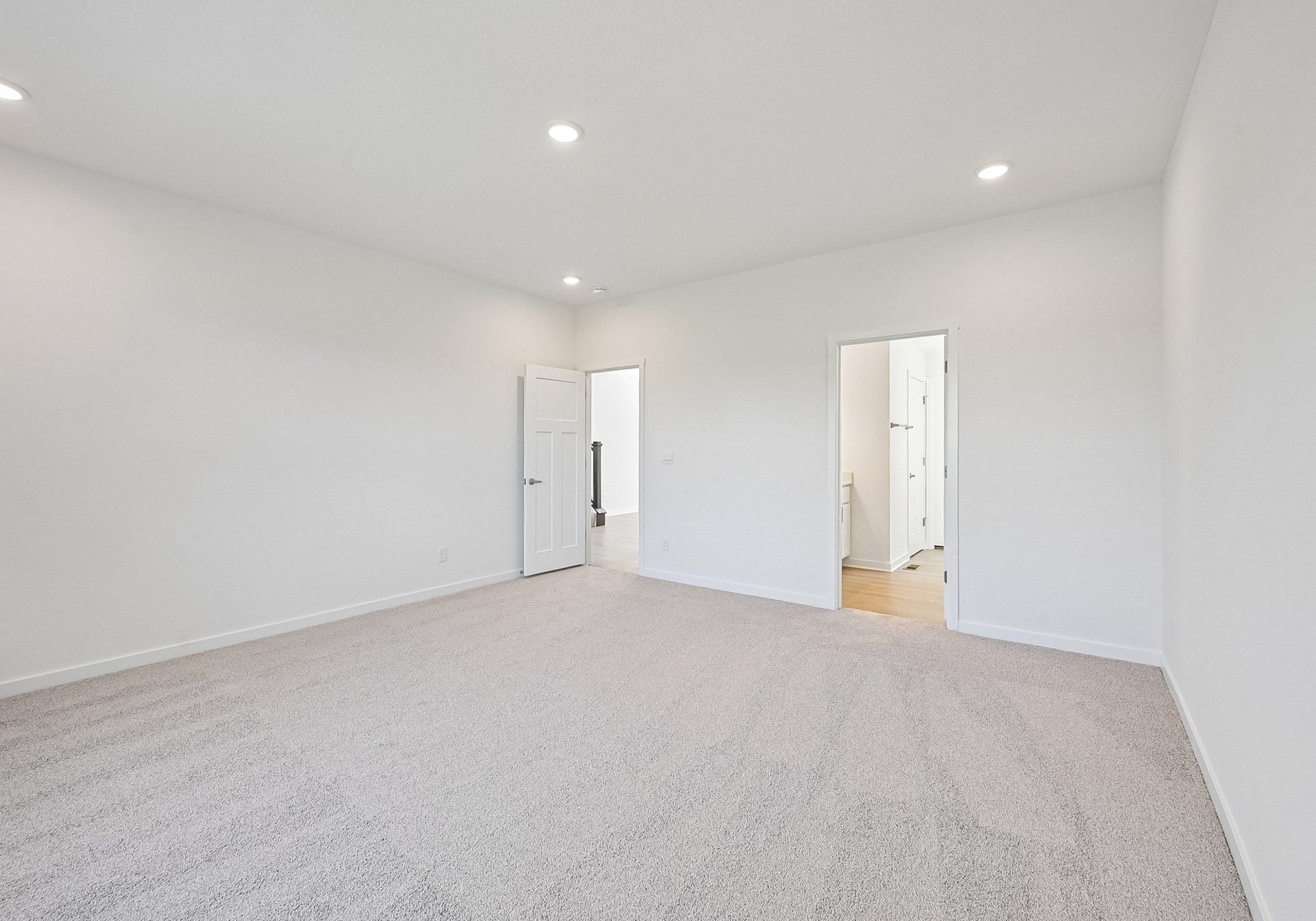 Spacious, empty room with light-colored walls and carpet, featuring an open doorway leading to another room.