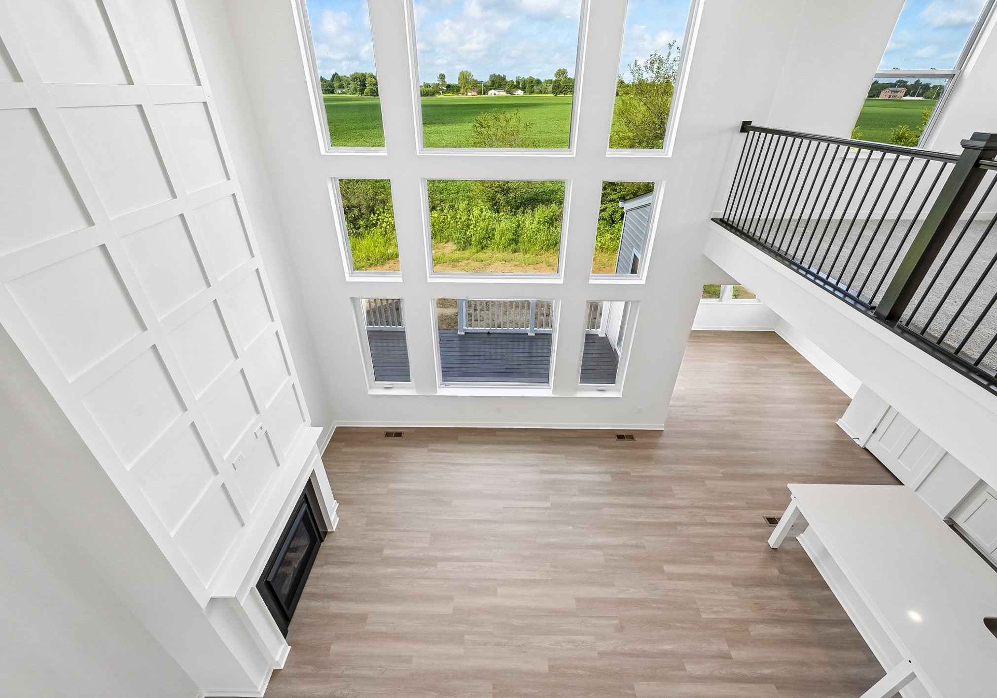 Bright, spacious living area featuring large windows, light wood flooring, and a white ceiling with a balcony above.