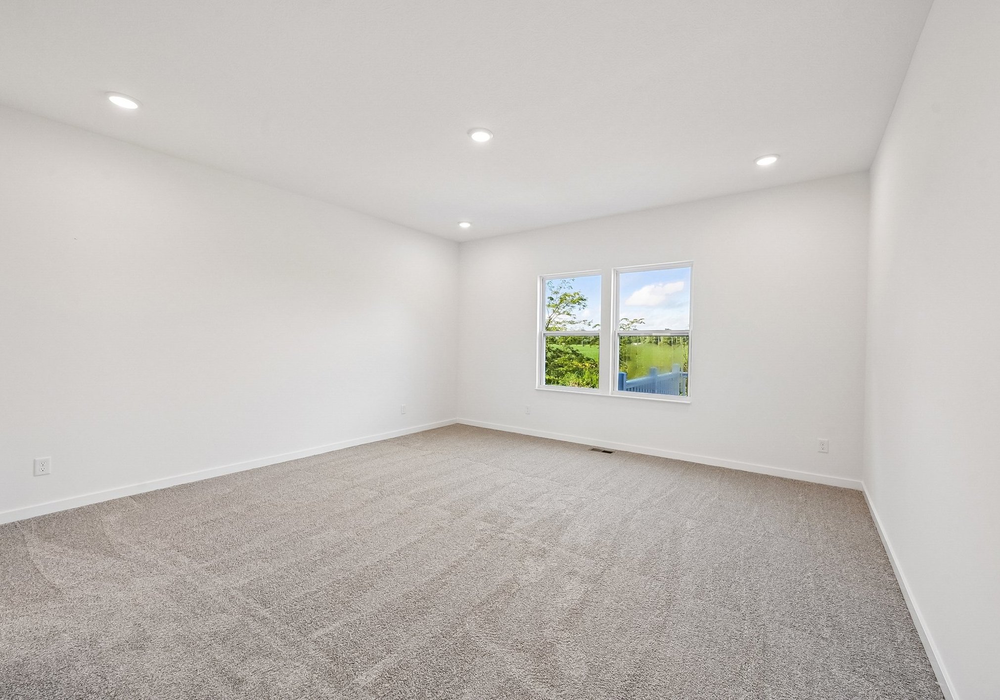 A spacious, empty room with light beige carpet and two windows letting in natural light.