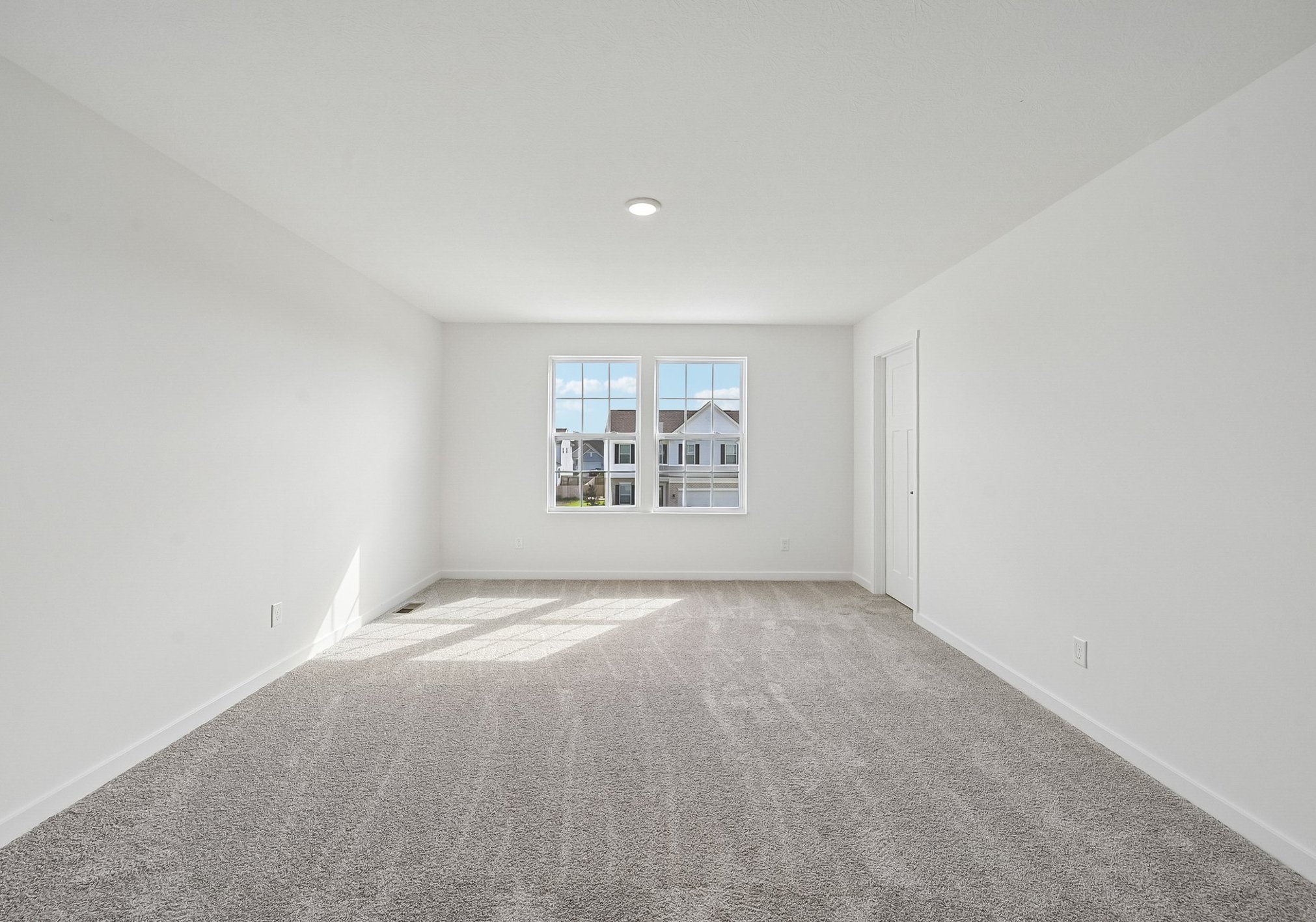 A spacious, empty room with gray carpet and large windows letting in natural light.