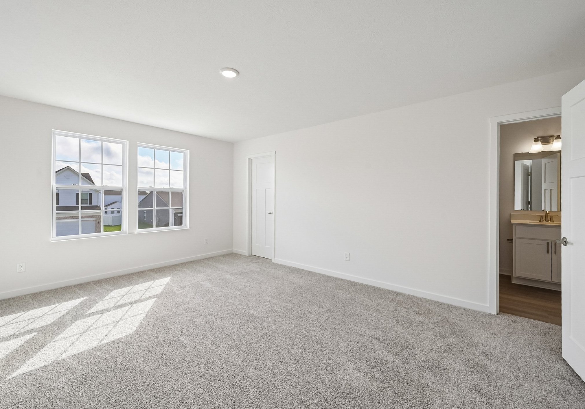Bright, empty room with light-colored walls, large windows allowing natural light, and a doorway leading to a bathroom.