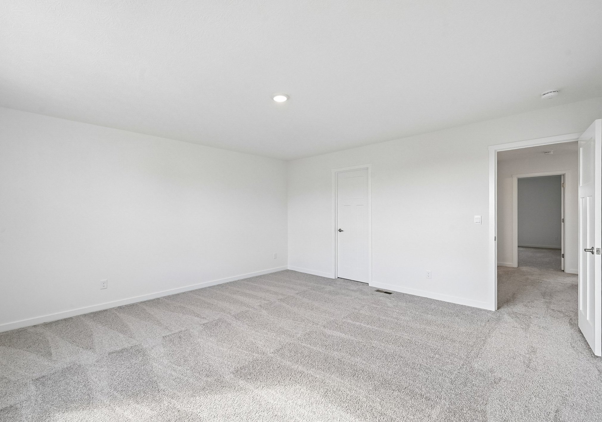 A spacious, empty room with light-colored walls and plush carpet, featuring a door leading to another room.