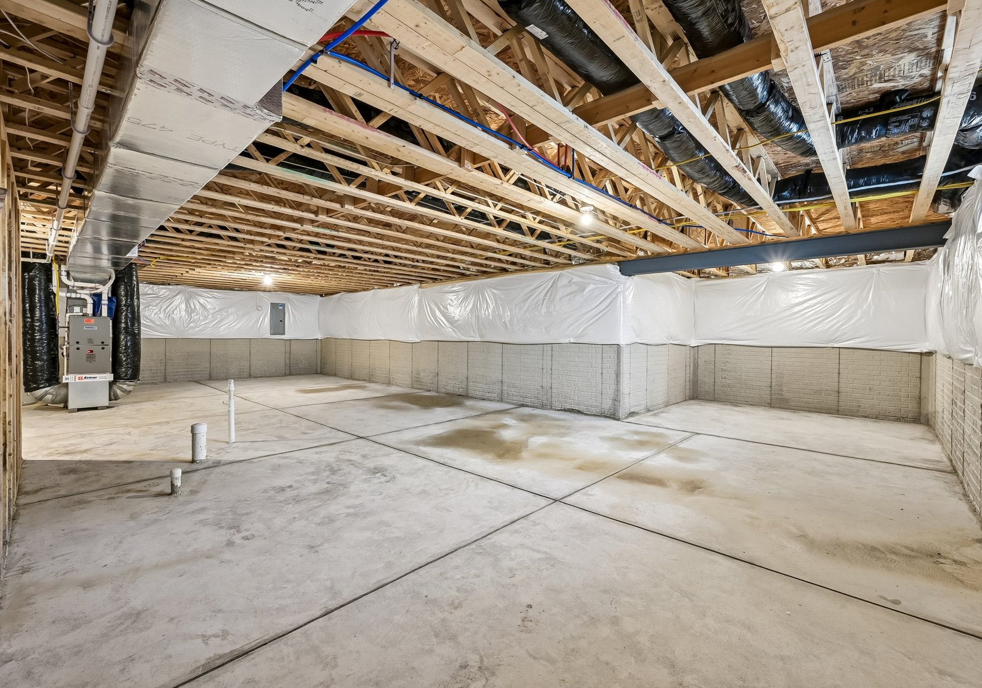 A spacious unfinished basement with exposed beams and concrete walls, featuring plastic sheeting and utility equipment.