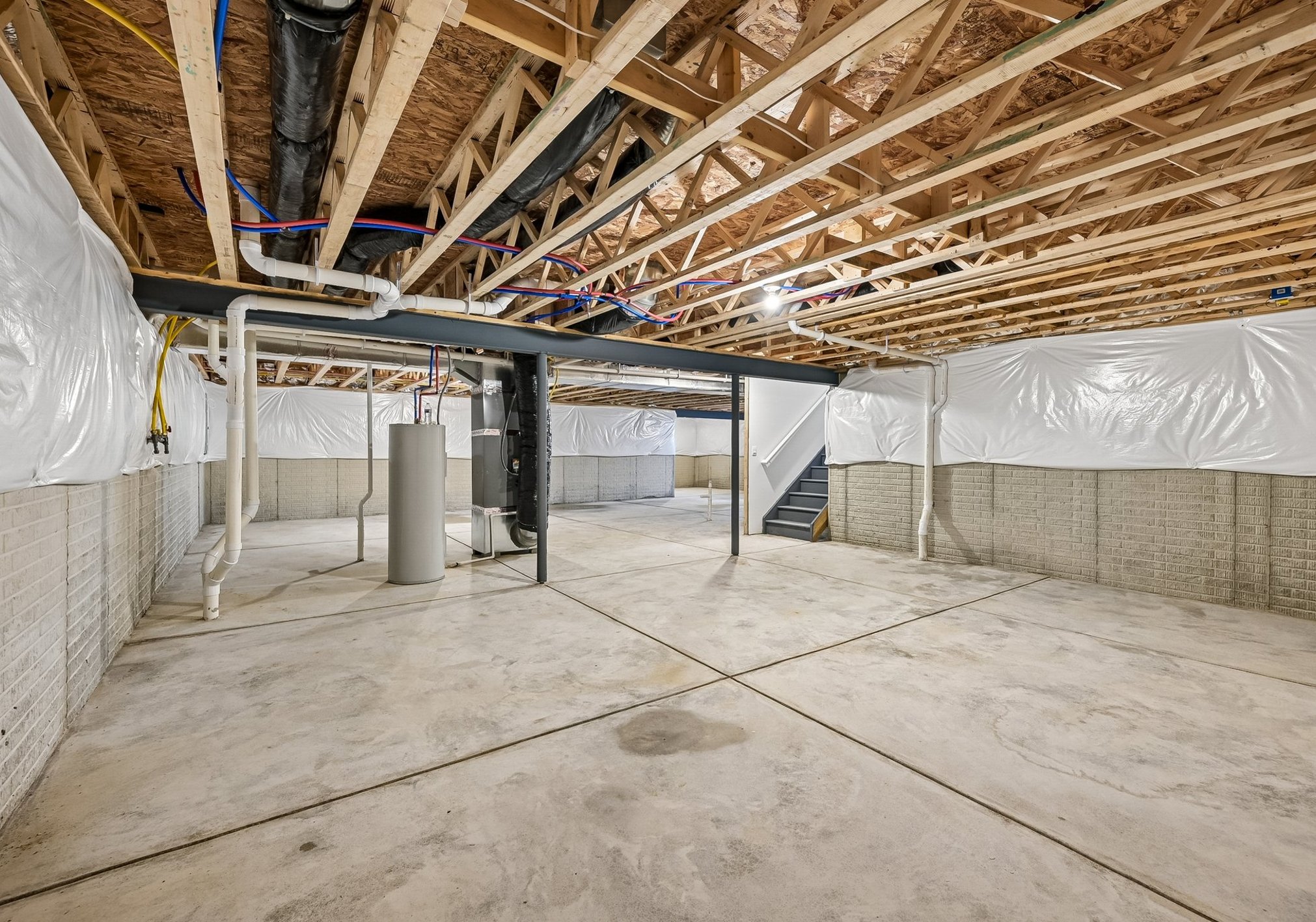 A spacious unfinished basement with exposed beams, concrete floors, and plastic-wrapped walls.