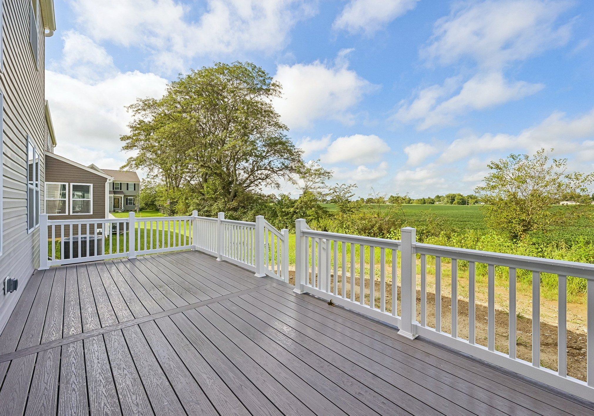 A wooden deck with white railings overlooks a green field and a tree under a partly cloudy blue sky.